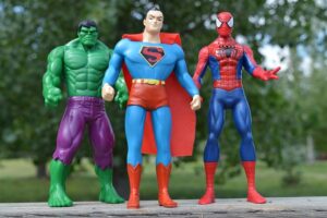 Three action figures of Hulk, Superman, and Spider-Man stand on a wooden surface outdoors with a blurred green background.