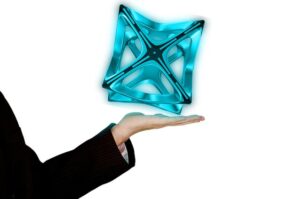Outstretched hand in a dark suit sleeve presents a blue, glassy, X-shaped geometric sculpture floating above the palm