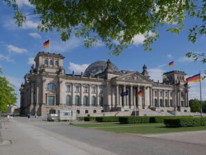 The Reichstag building in Berlin with a glass dome, flags on flagpoles, and a manicured lawn under a blue sky.