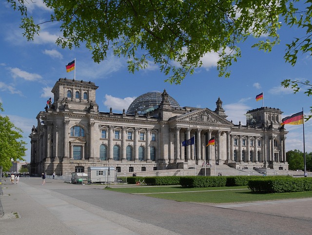 The Reichstag building in Berlin with a glass dome, flags on flagpoles, and a manicured lawn under a blue sky.