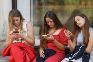 Three young women sitting on a concrete step, each looking at their smartphone screens outside a building.
