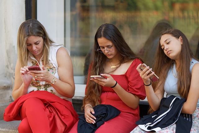 Three young women sitting on a concrete step, each looking at their smartphone screens outside a building.