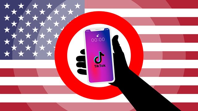 Silhouette hand holding a smartphone showing the TikTok logo inside a red prohibition circle, with a US flag backdrop.