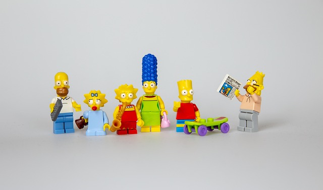 Row of LEGO Simpsons minifigures lined up on a gray surface, including Homer, Marge, Lisa, Maggie, Bart, and a figure on the right reading a newspaper.