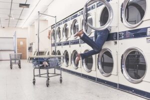 Person's leg sticks out of a front-loading dryer in a laundromat, with empty carts nearby and row of machines along the wall.