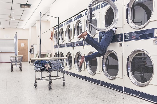 Person's leg sticks out of a front-loading dryer in a laundromat, with empty carts nearby and row of machines along the wall.