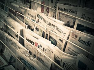 Piled German newspapers in a rack with front pages visible, including titles like Neue Ruhr Zeitung and Welt, in a newspaper stand setting.