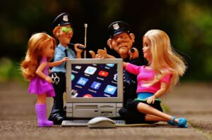 Toy dolls and police figure characters gather around a small computer monitor displaying social media icons (LinkedIn, Facebook, Twitter).