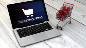 Laptop shows an online shopping logo while a small shopping cart with gift boxes sits on a light wooden floor, conveying e-commerce shopping.