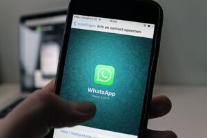 Smartphone held in hand displaying the WhatsApp splash screen with the green logo on a teal patterned background, in app launch view.